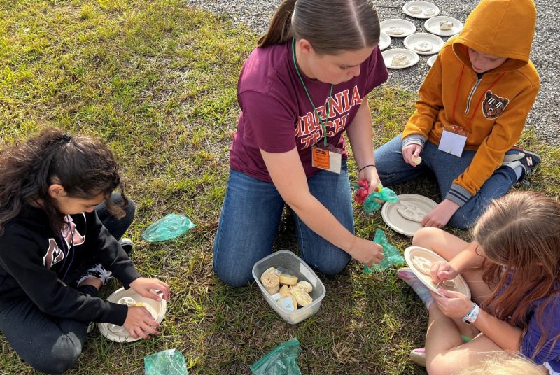 Five children and a college student sit in a circle on the grass. The students are holding or touch paper plates with round, flat pieces of clay, each with an animal footprint.