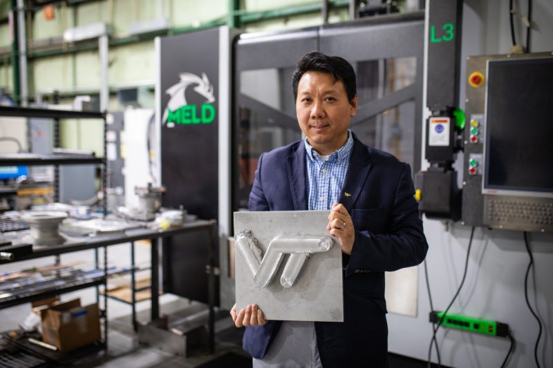 A man in a navy blazer and blue checkered shirt stands in front of a MELD 3D metal printing machine, holding a square metal plate featuring a raised Virginia Tech logo made from metal deposition.