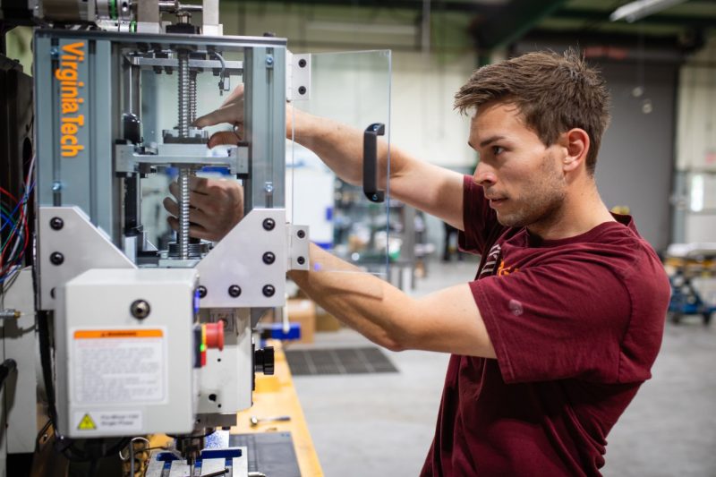 A graduate student in a Virginia Tech shirt tinkers with a small additive friction stir deposition machine in a large lab.