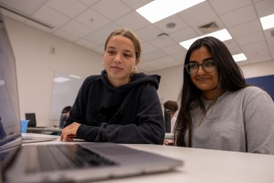 Two female students, one in a blue sweatshirt and one in a gray, look at a computer screen in class.