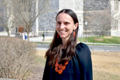 Wearing a blue pullover adorned with an orange-colored necklace, Assistant Professor Lauren Childs poses near Torgersen Hall. 