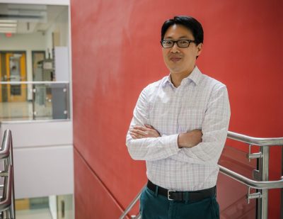Vincent Wang, associate professor in biomedical engineering and mechanics, stands in the foyer of Kelly Hall with a red background. Photo by Spencer Roberts of Virginia Tech
