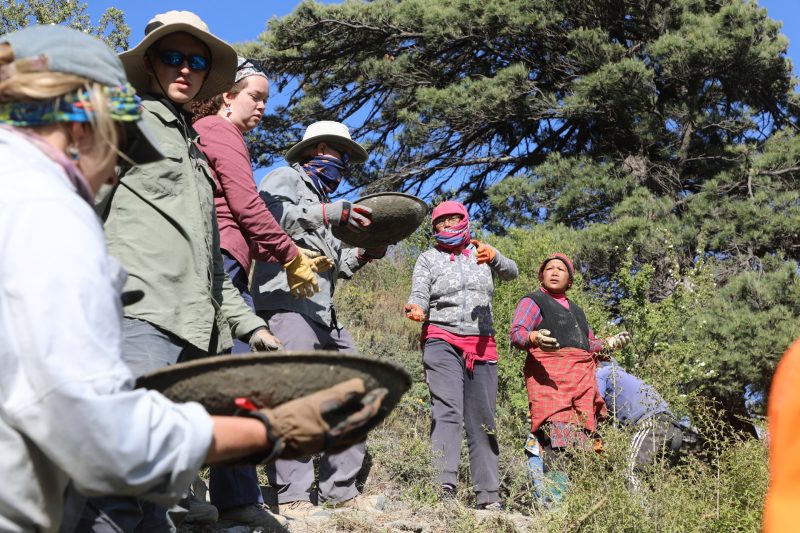 A row of seven people in sun-blocking clothing pass pans of concrete up a rocky, treed hillside.