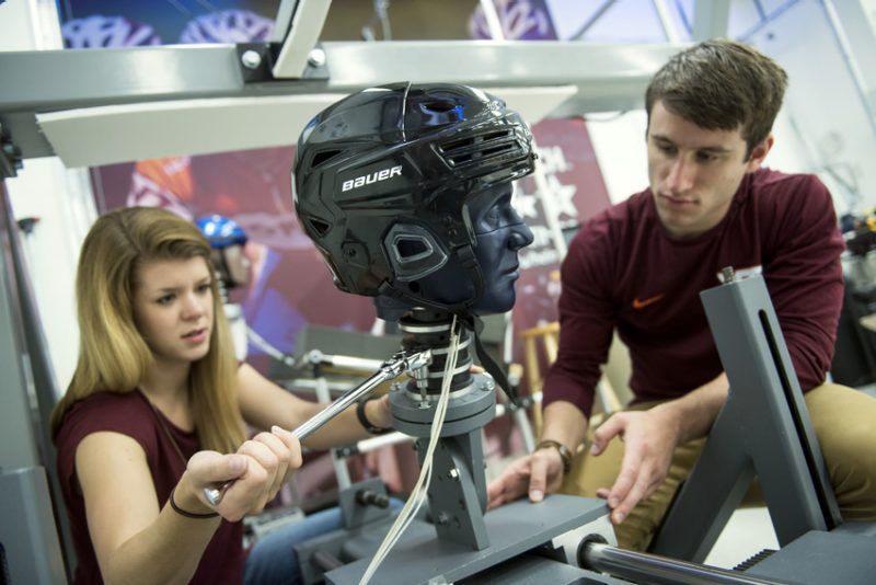 Two researchers working within the Helmet Lab, the researcher on left is operating a ratchet handle while the researcher on the right steadies the frame of an experiment.
